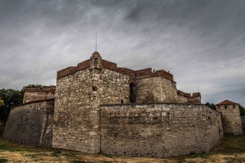 Панорамен изглед към масивните каменни зидове и отбранителни кули на крепостта „Баба Вида“ под тежко облачно небе.