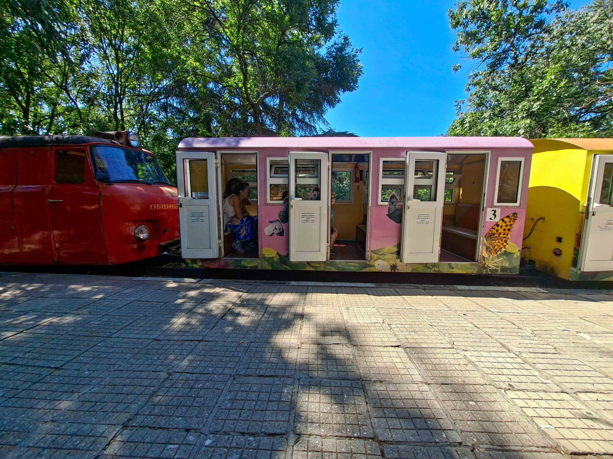 Розов wagon от Детската железница в Пловдив с отворени врати, спрял на перона сред сенките на дърветата.